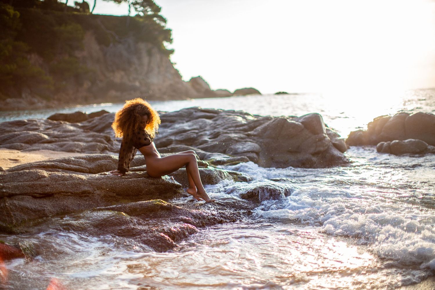 Woman with curly hair sitting on rocky shoreline at sunset, waves washing over rocks at a scenic coastal beach.