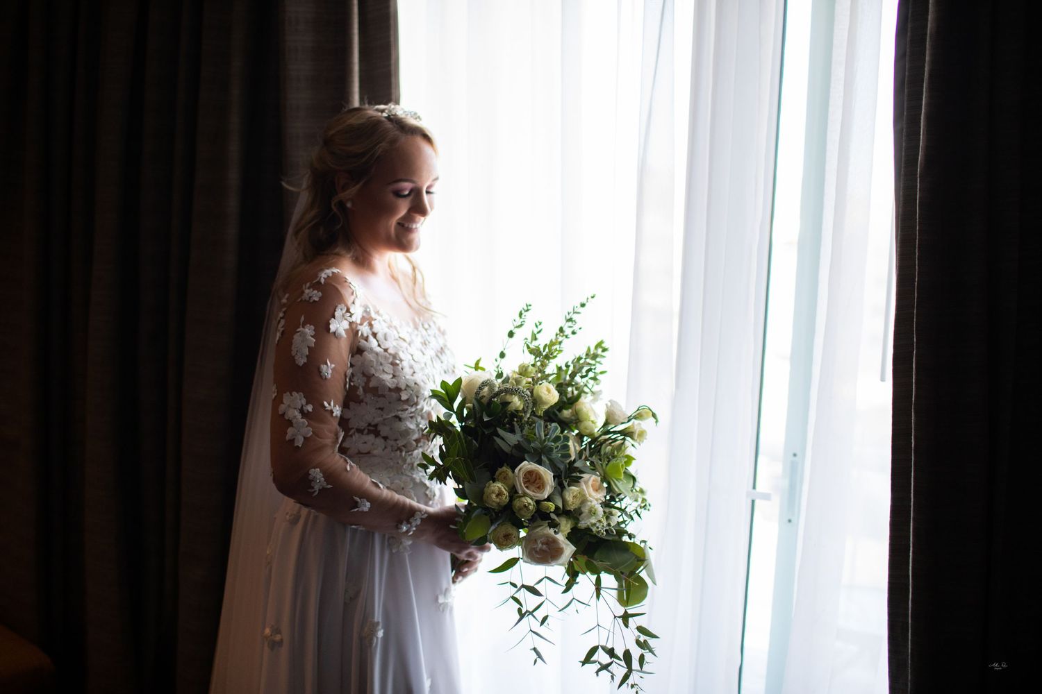 Elegant bride stands by a window in soft light, holding a lush white and green wedding bouquet.
