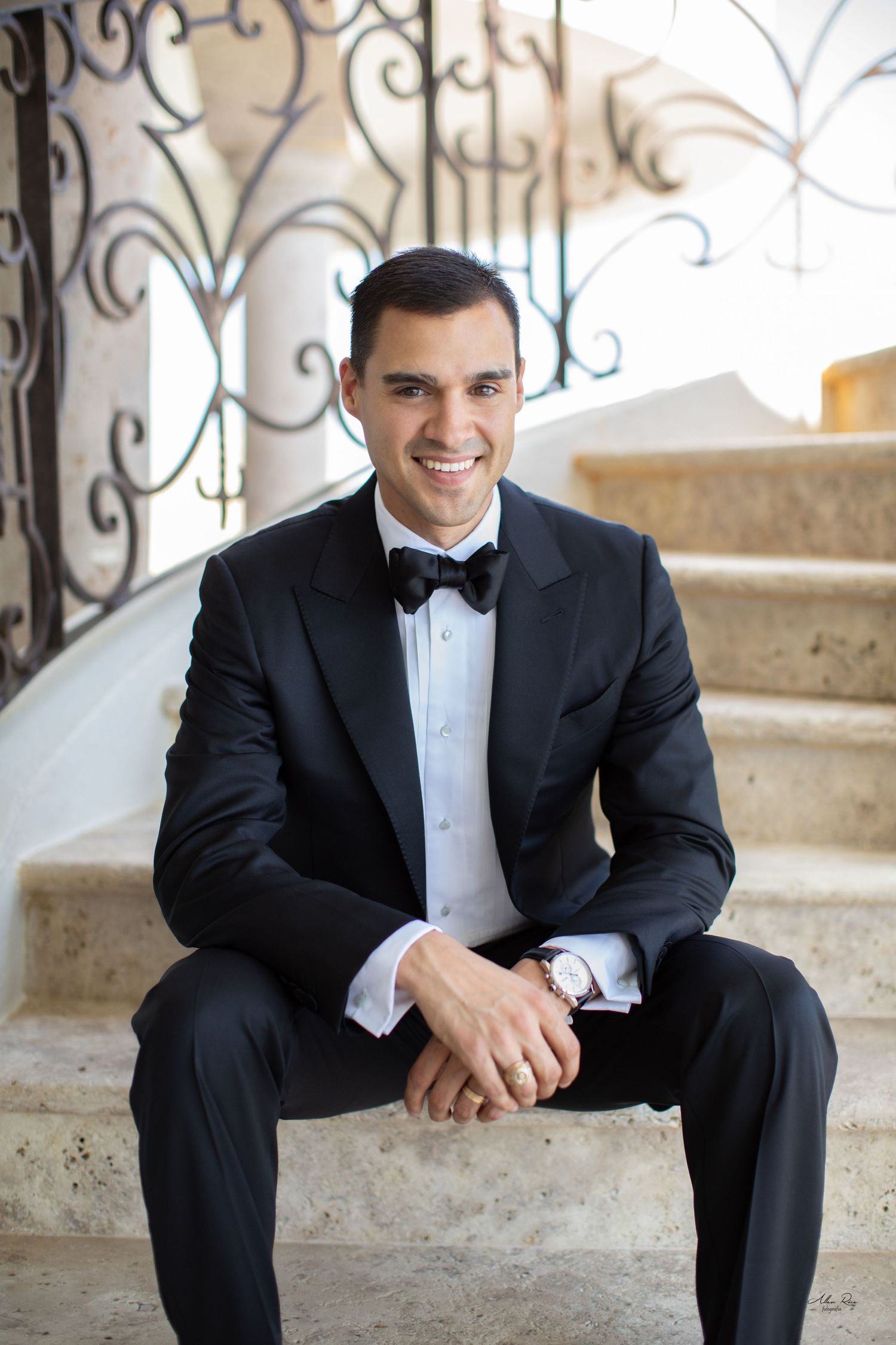 Handsome groom in black tuxedo with bow tie sits smiling on marble staircase with decorative iron railing.