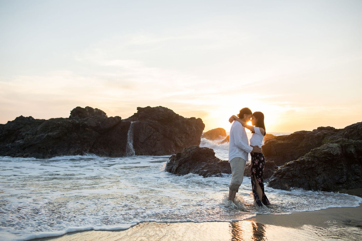 Couple embracing in ocean waves on rocky beach during breathtaking sunset.