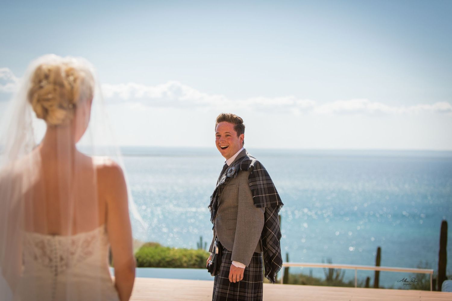 Groom in a tartan kilt smiling at his bride with a stunning ocean backdrop.