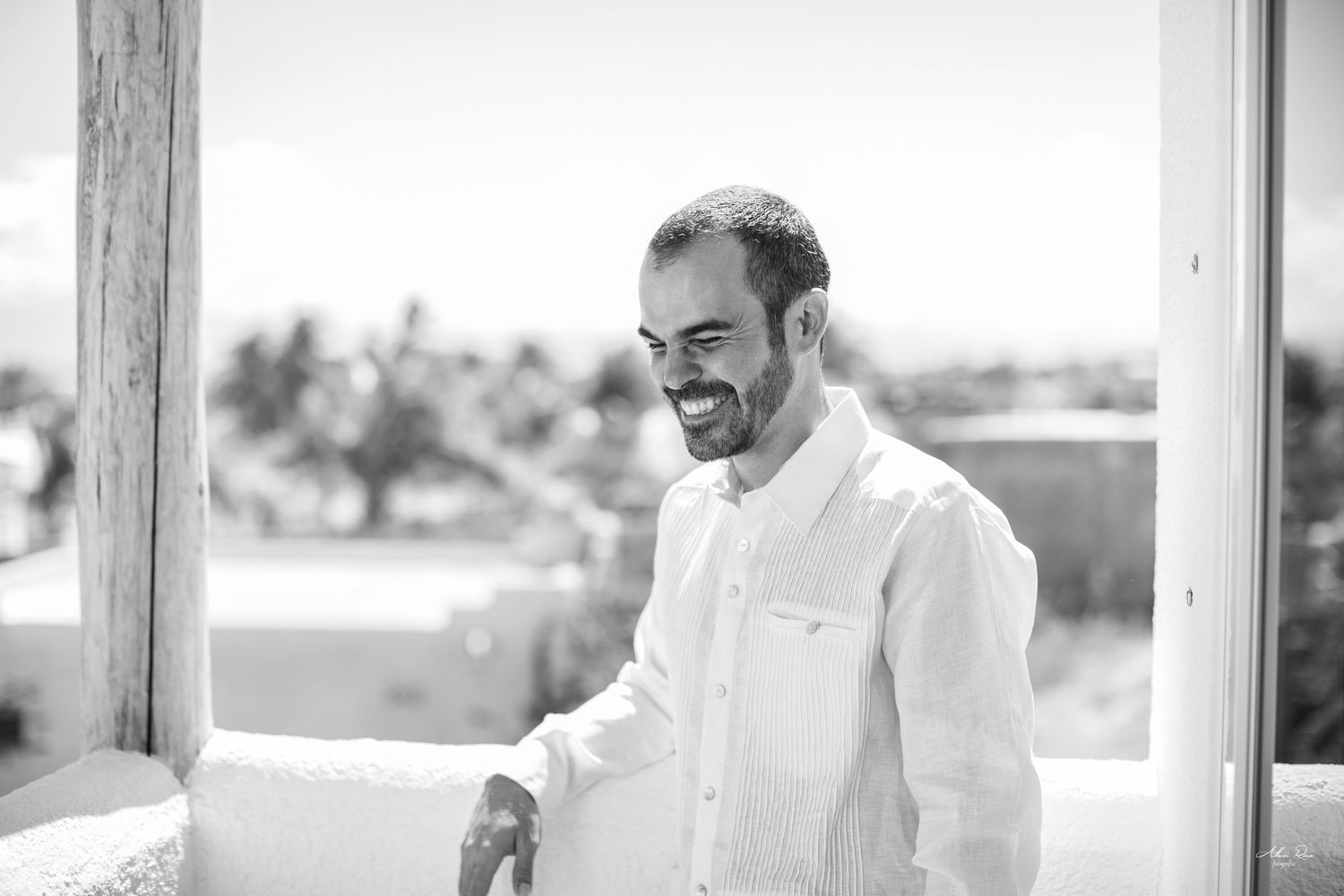 Smiling groom in white linen shirt laughing outdoors on sunny wedding day in black and white.