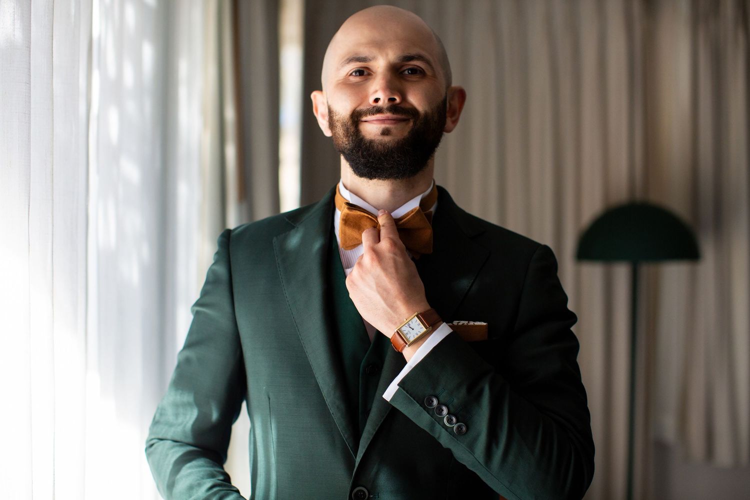 Bald groom with beard adjusting orange bow tie wearing elegant dark green suit indoors.