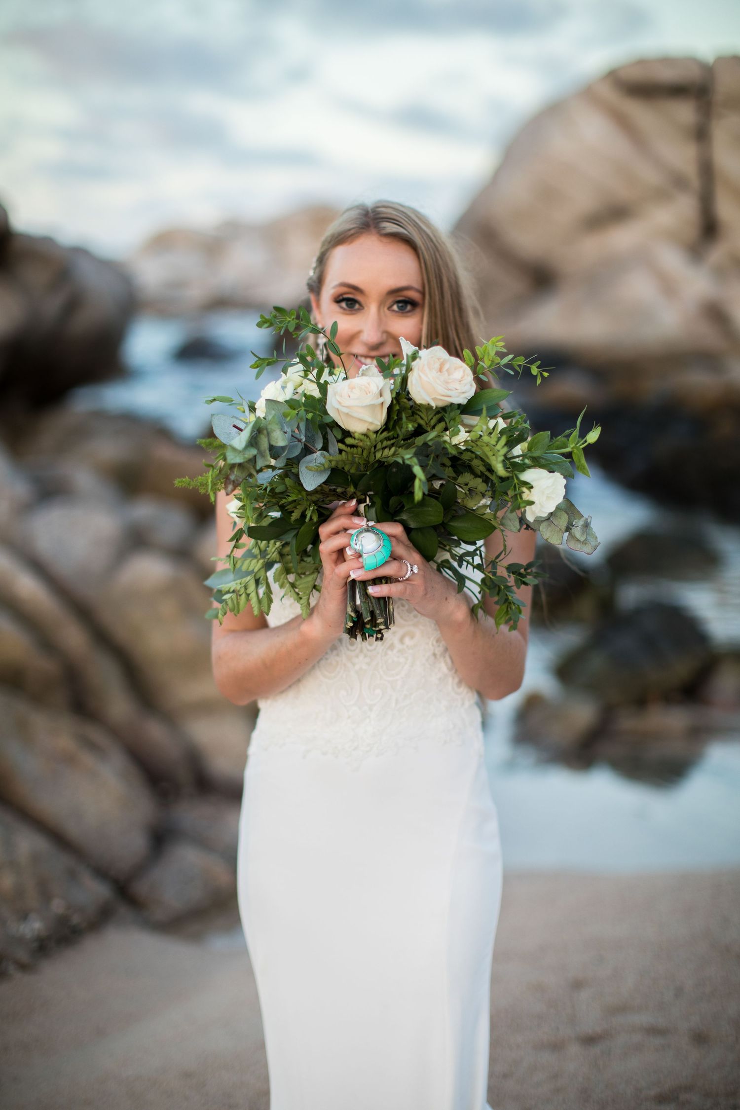 Bride in white lace gown holding a lush bouquet of white roses and greenery at the beach.