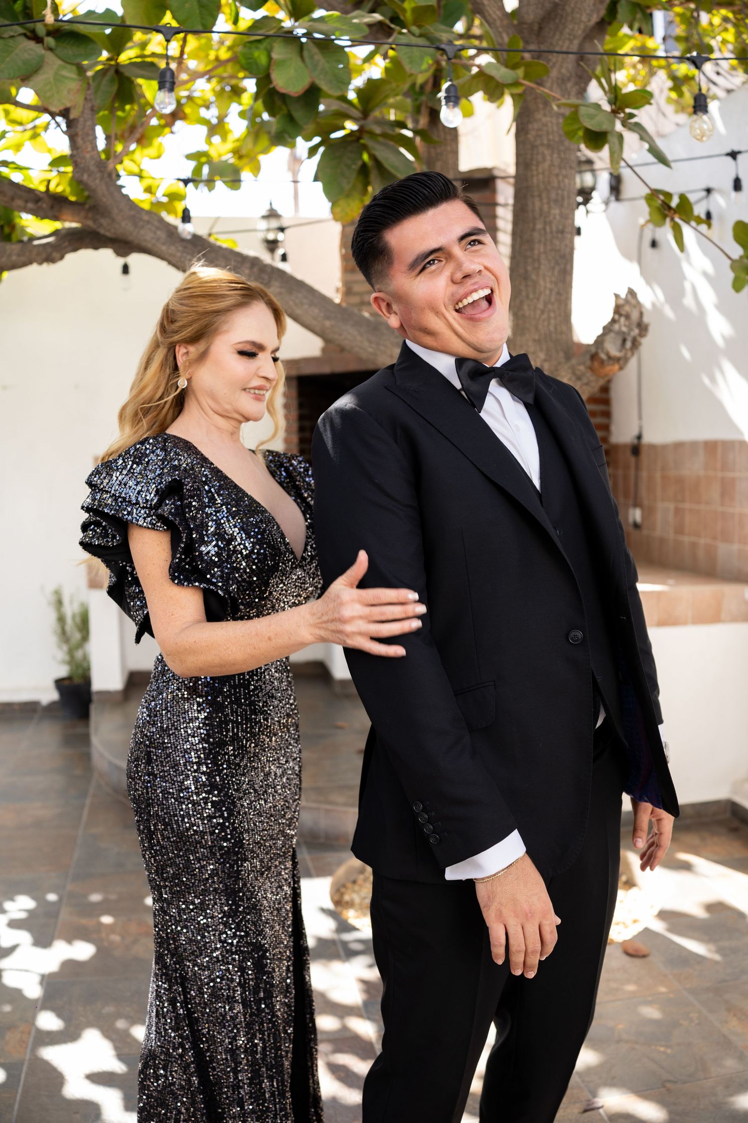 Laughing groom in black tuxedo walks with elegant bride in sequin gown under a sunlit tree.