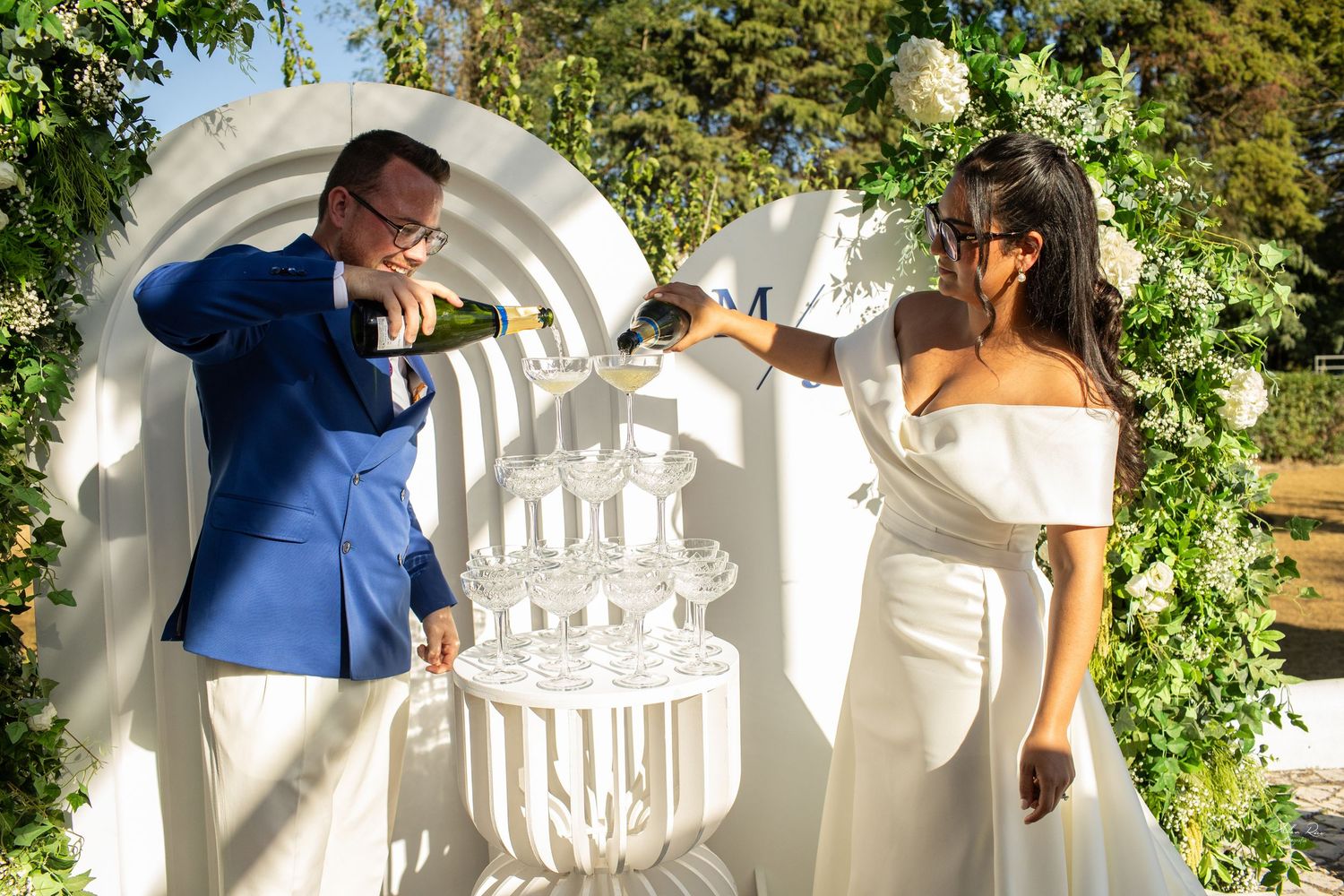 Bride and groom pour champagne into a glass tower at their elegant outdoor wedding celebration.