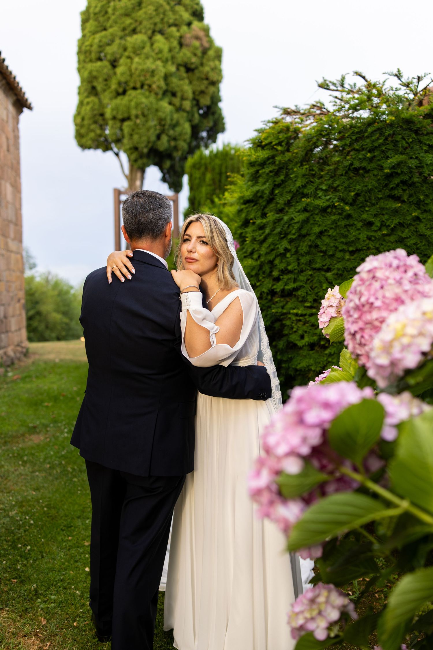 Bride in white veil embraces groom in garden with pink hydrangeas in foreground.