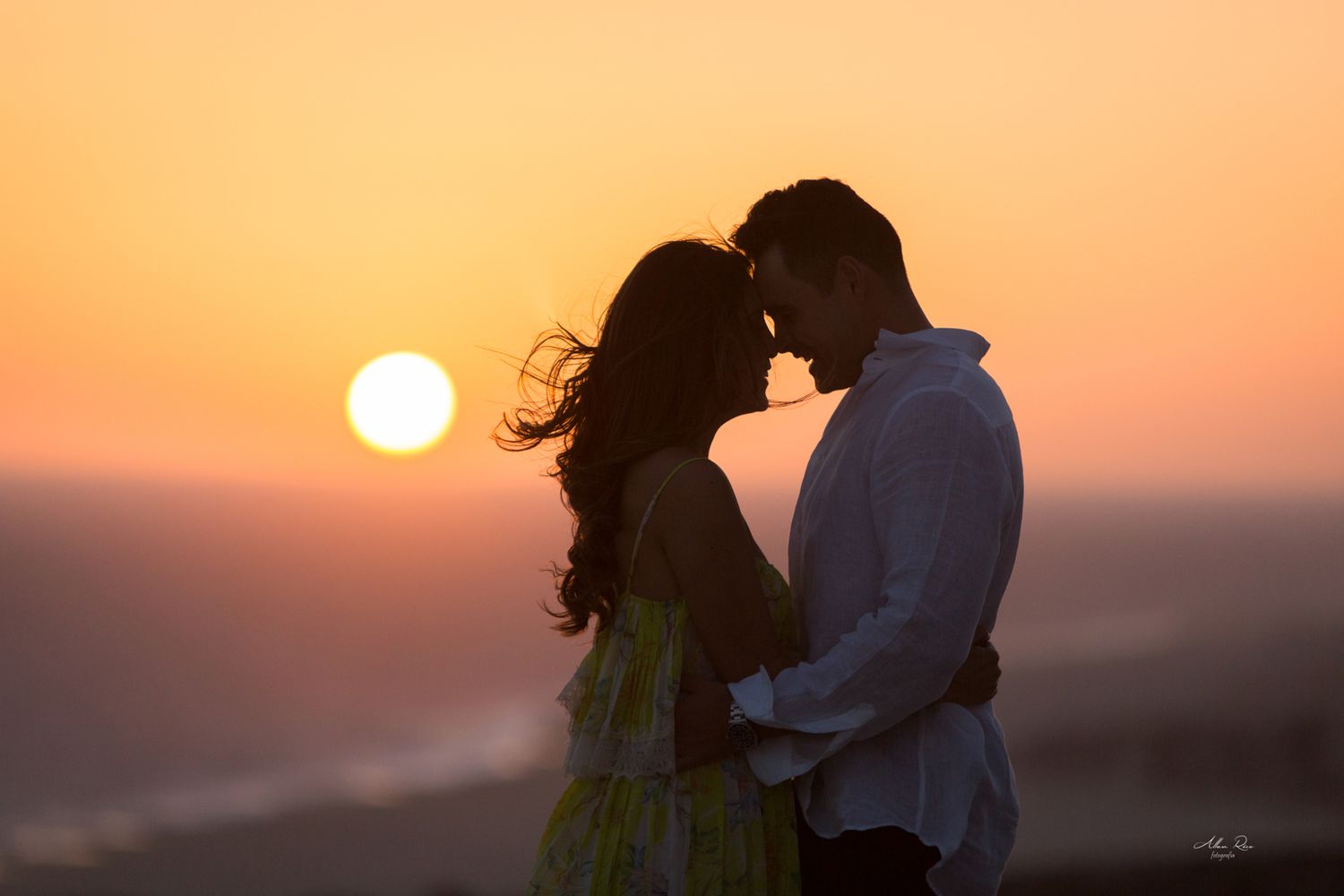 Couple sharing an intimate moment with foreheads touching against a glowing orange sunset.