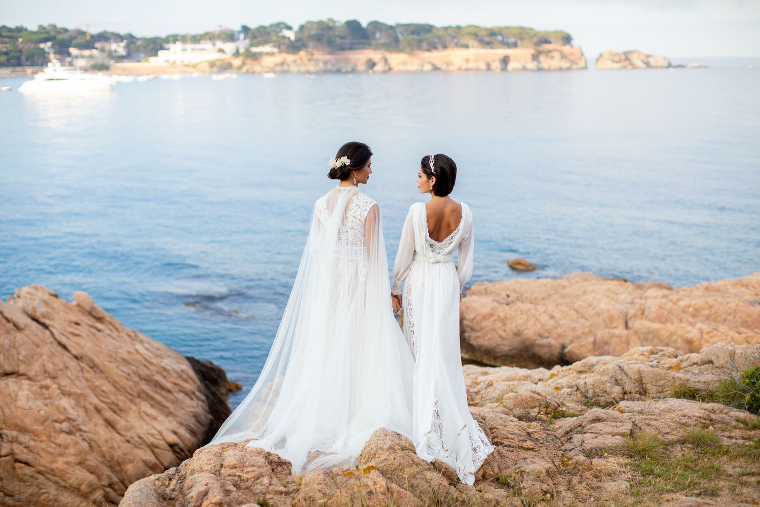 Two brides in elegant white wedding gowns stand back-to-back on rocky cliffs overlooking the sea.