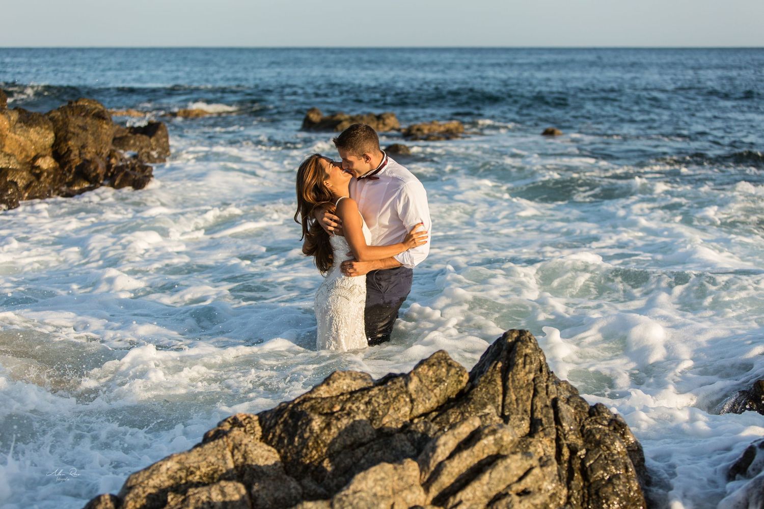 Couple embraces romantically amid crashing ocean waves on rocky coastline in bright sunny conditions.
