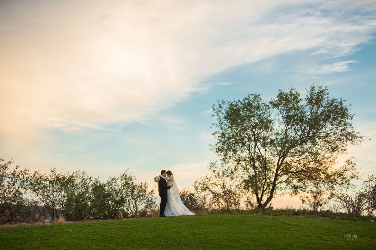 Bride and groom embrace on a lush green lawn beneath a dramatic golden sunset sky.
