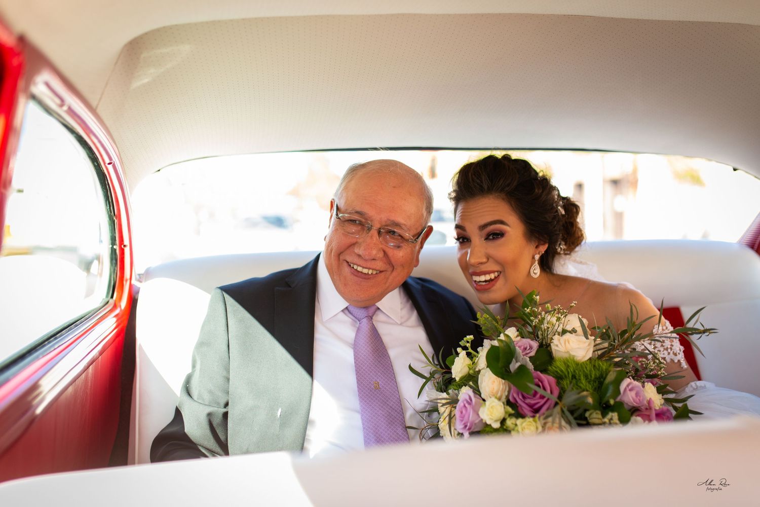 Smiling bride and older man posing together in backseat of a classic red and white vintage car.