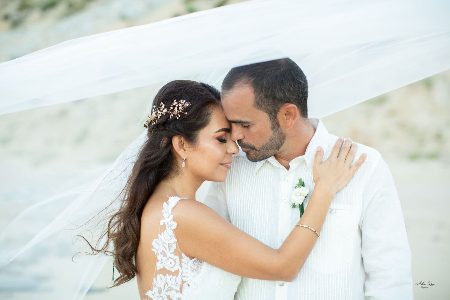 Bride and groom share an intimate moment under flowing white veil on a serene sandy beach.