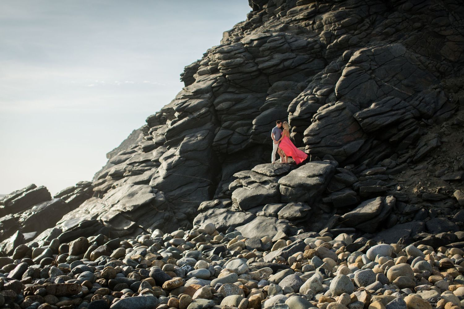Couple in formal wear climbing rocky coastal cliffs, woman in flowing red dress.