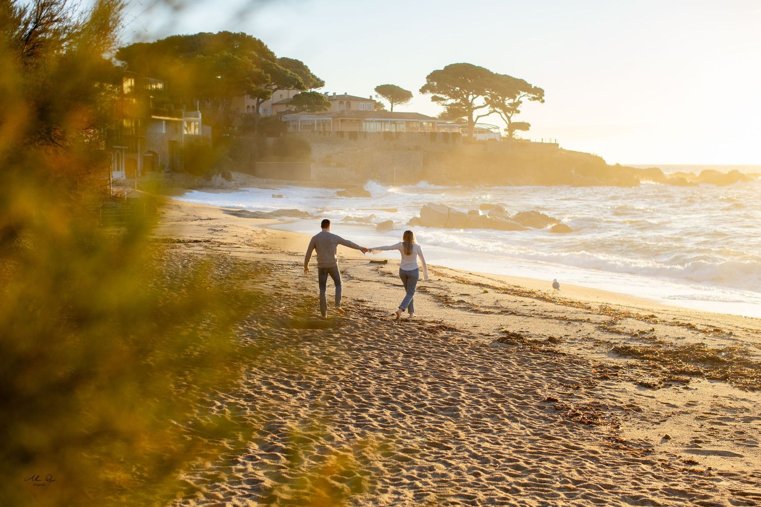 Couple walking away hand in hand along a golden sandy beach at sunset with waves crashing nearby.