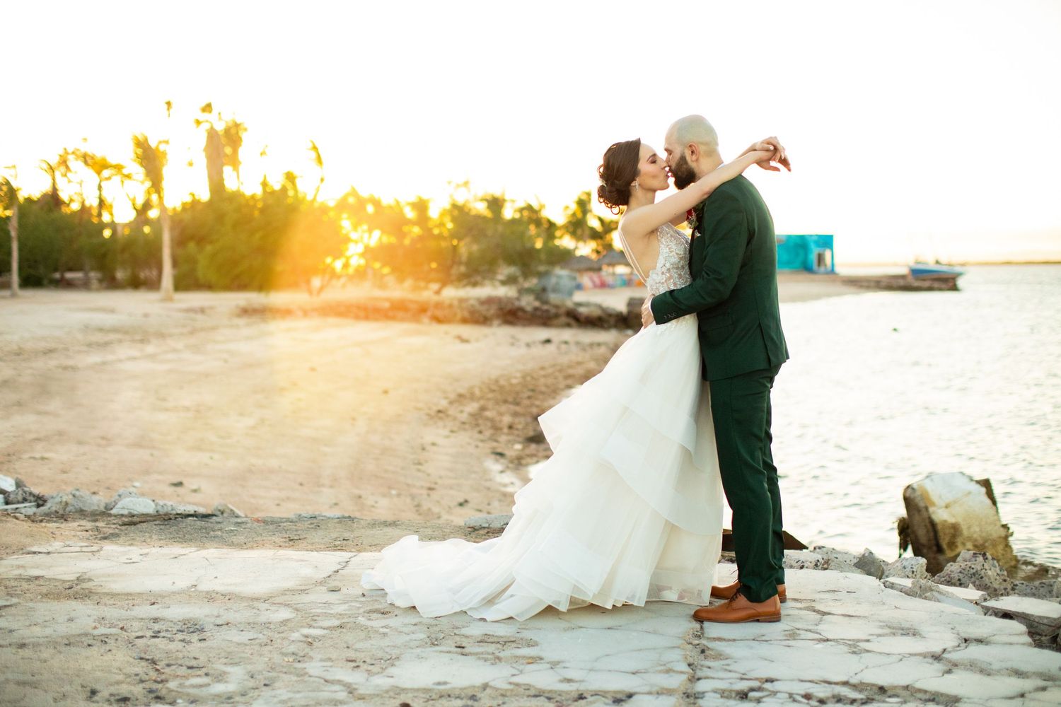 Bride and groom sharing romantic kiss at sunset on rocky waterfront in wedding attire.
