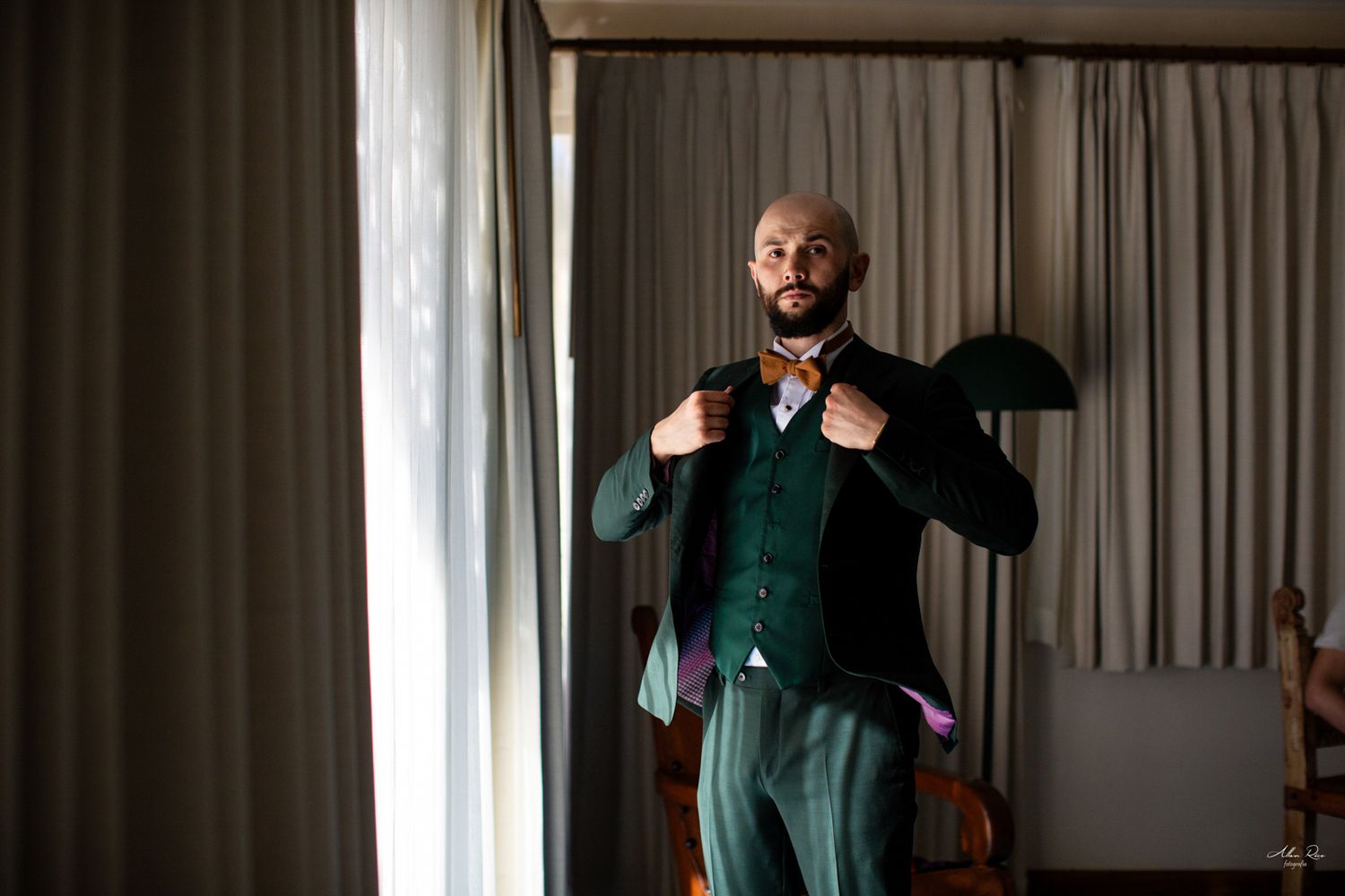 Bald groom adjusting bow tie while getting dressed in elegant dark green suit by window.