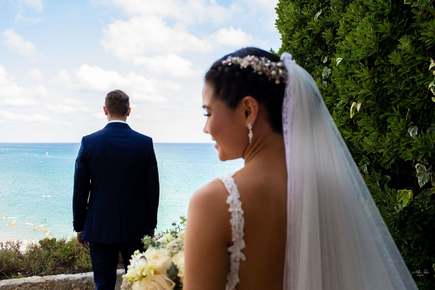 Bride in white lace gown and veil smiles as groom in navy suit gazes at turquoise ocean at beach wedding.
