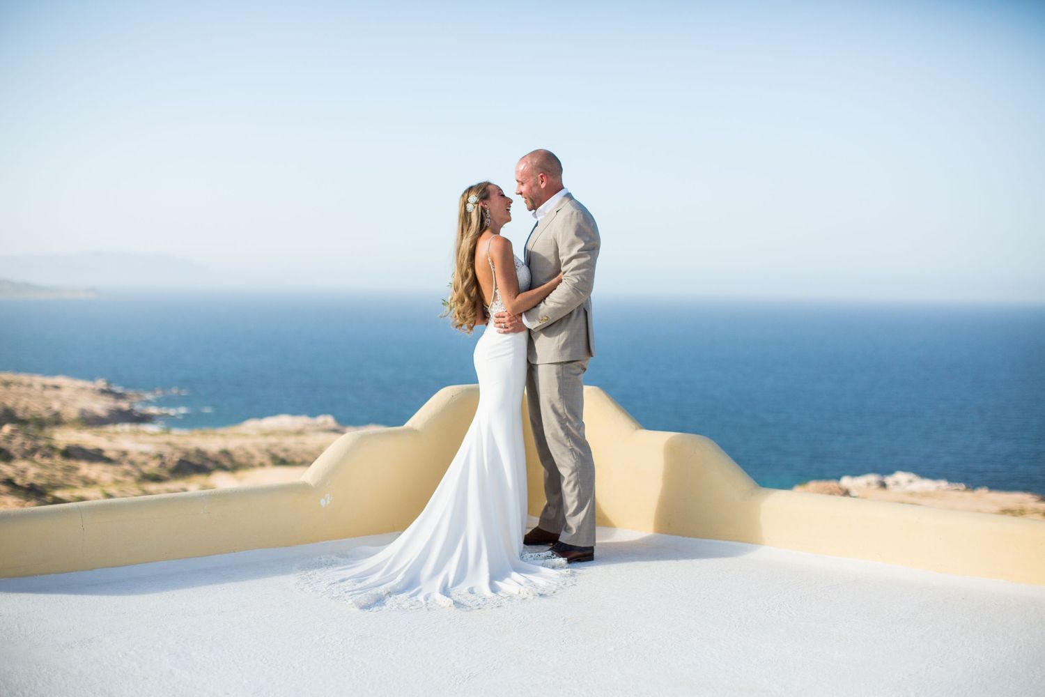 Bride and groom share a romantic kiss on a white terrace overlooking the stunning blue Mediterranean Sea.