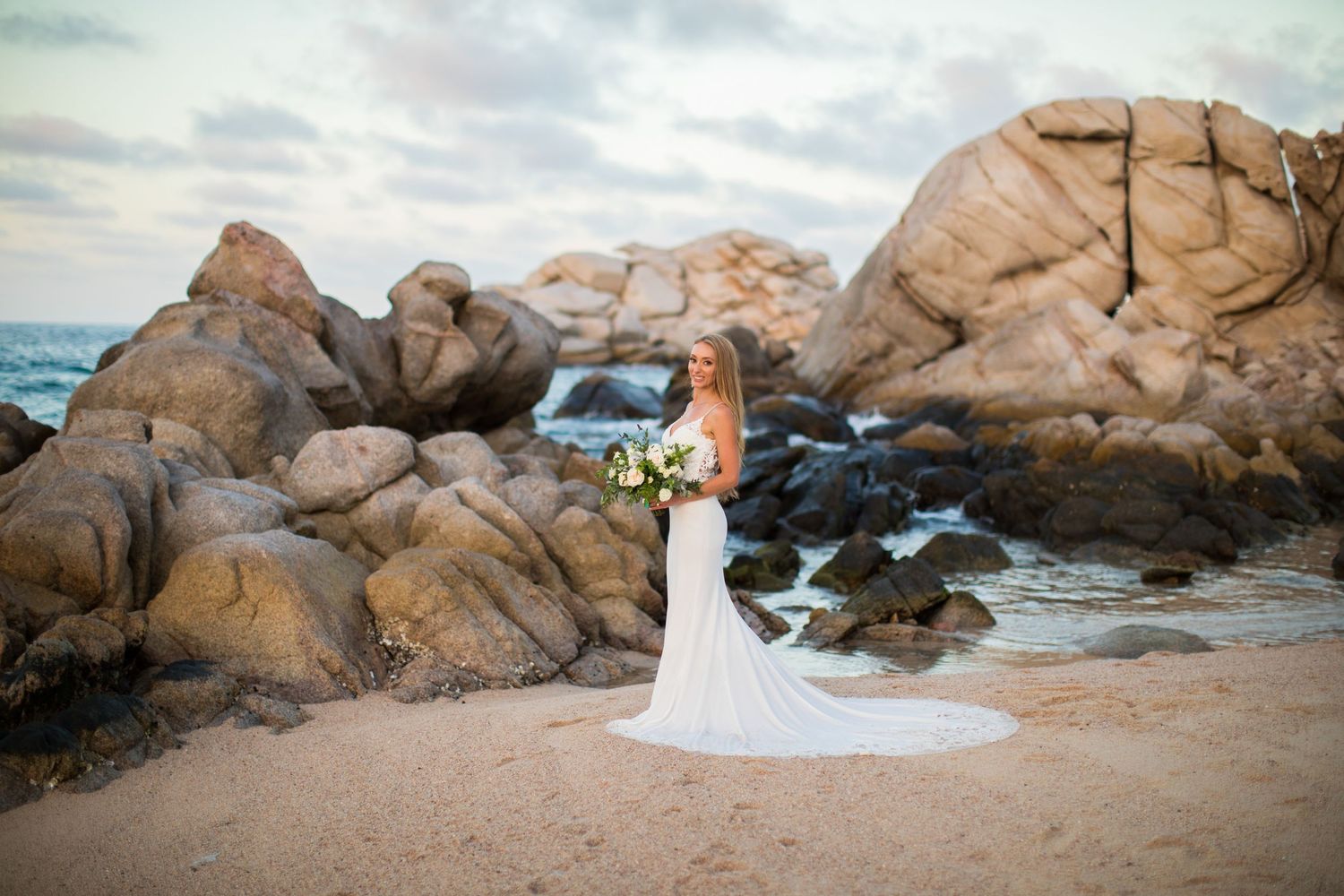 Elegant bride in fitted white gown poses on sandy beach surrounded by large granite boulders.
