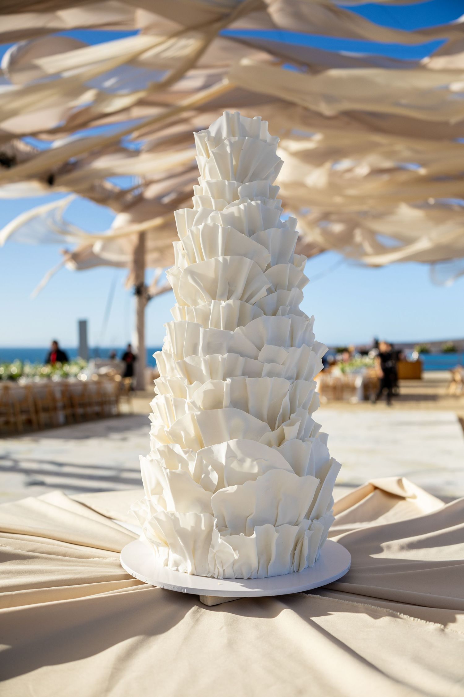 Tall sculptural white wedding cake with layered petal design at outdoor seaside venue.