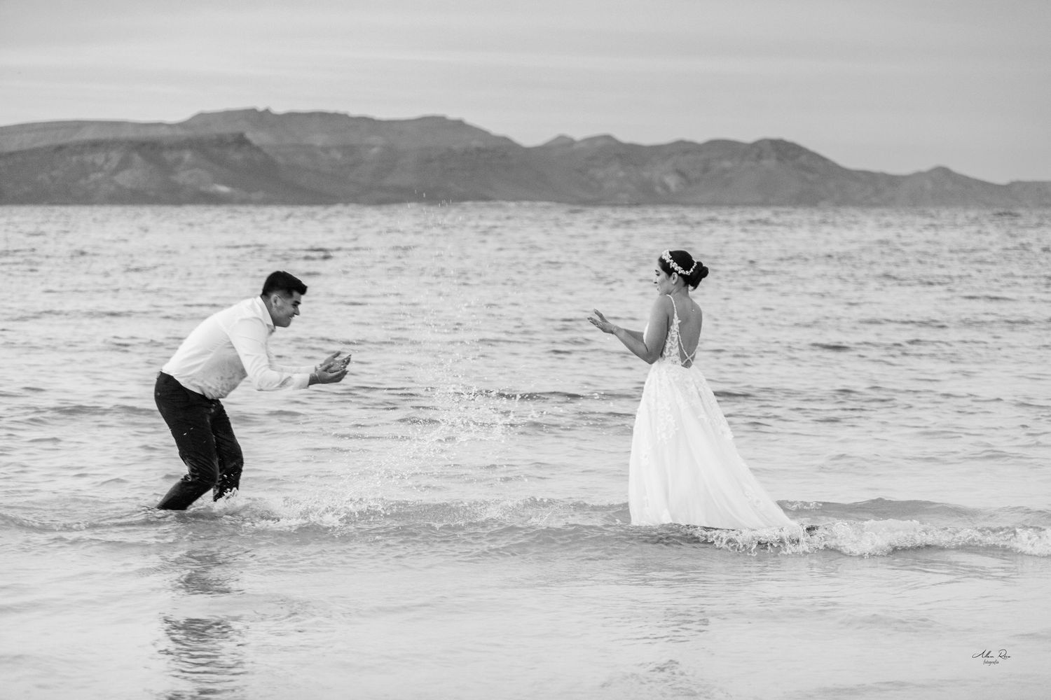 Black and white photo of bride and groom playfully posing in ocean waves with mountain backdrop.