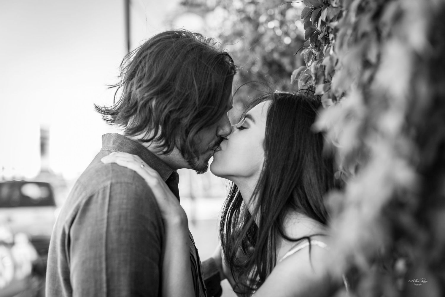 Couple shares a passionate kiss outdoors in a black and white engagement photo surrounded by lush foliage.
