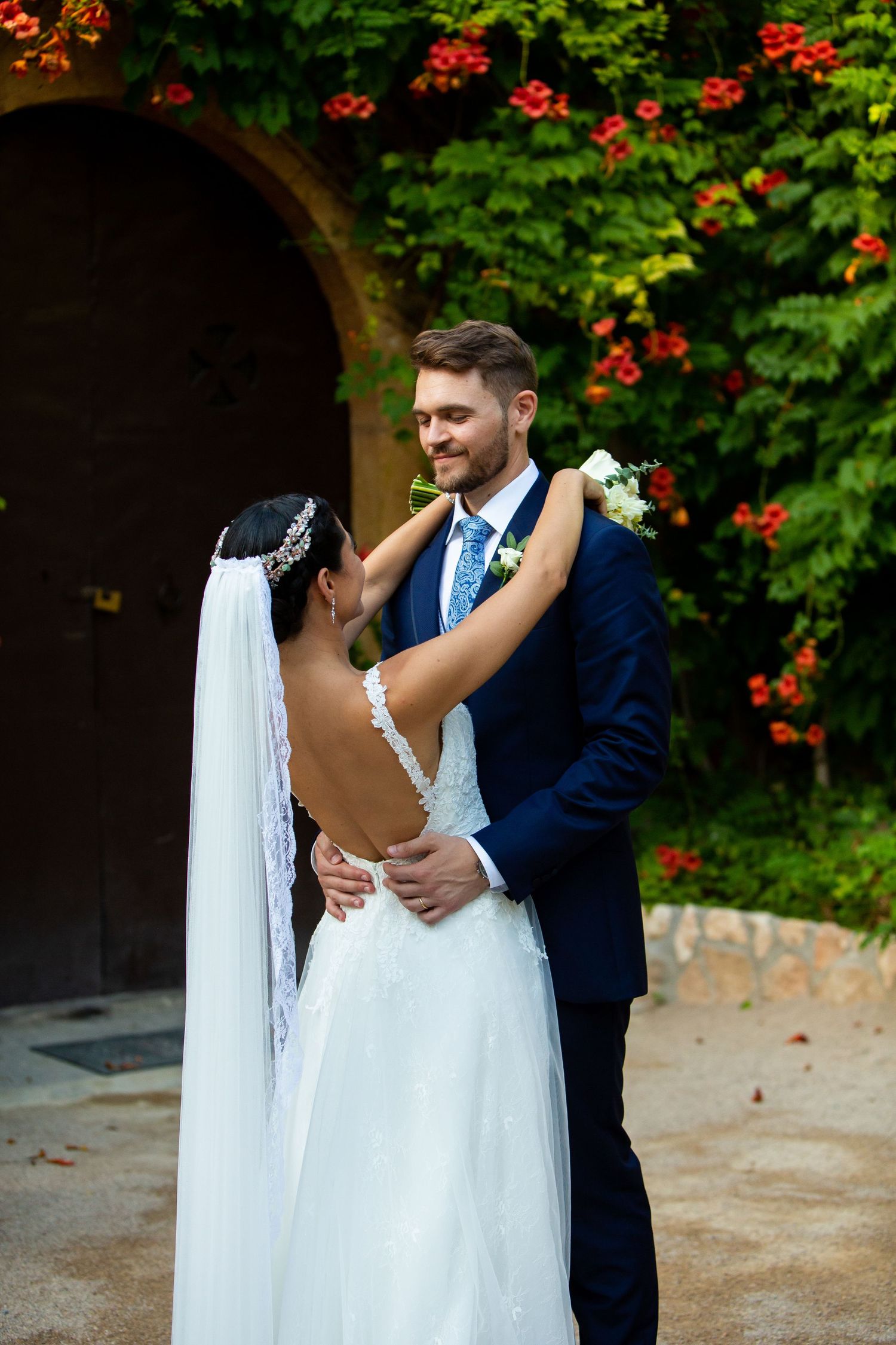 Groom smiling as bride embraces him near floral arch at outdoor wedding venue.