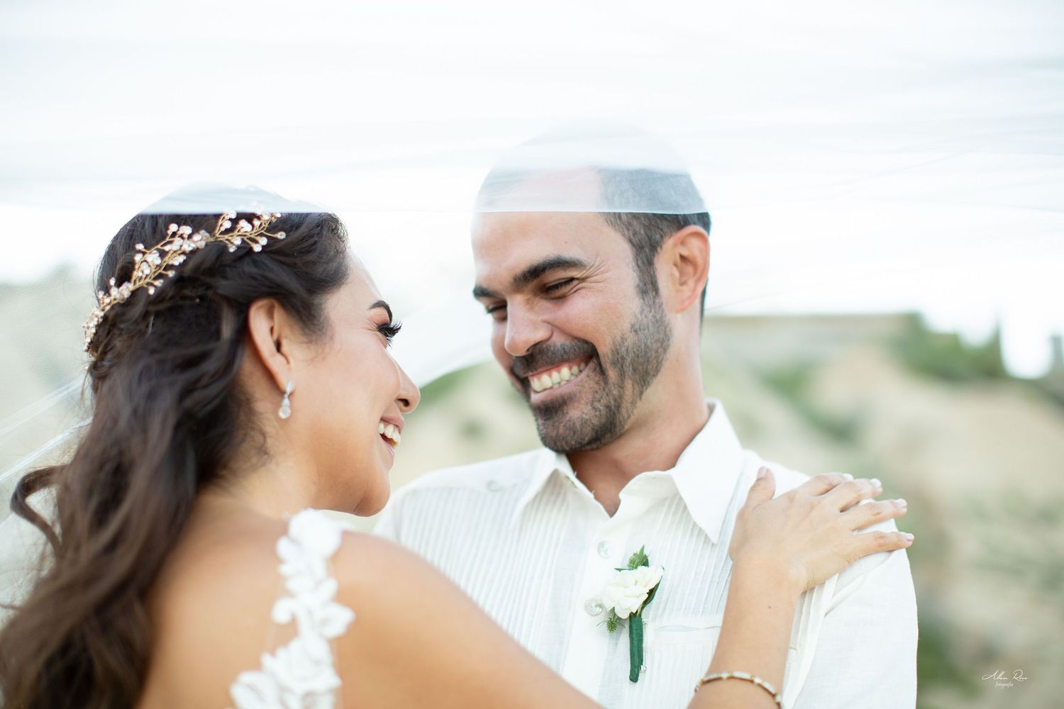 Smiling groom in white kippah and boutonniere gazes lovingly at bride during outdoor beach wedding.