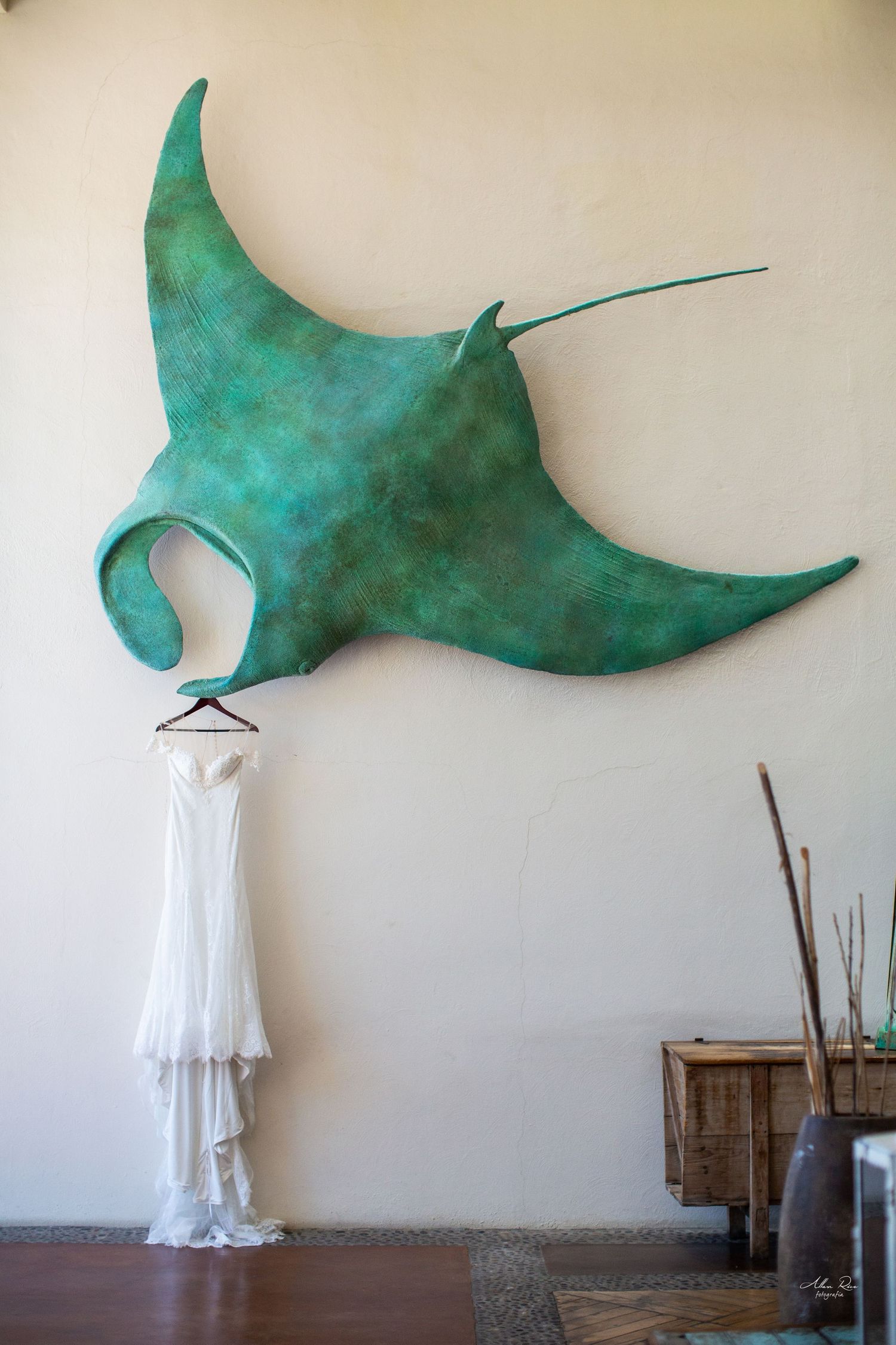 White wedding dress hanging on a hanger beneath a large green manta ray wall sculpture in an elegant venue.