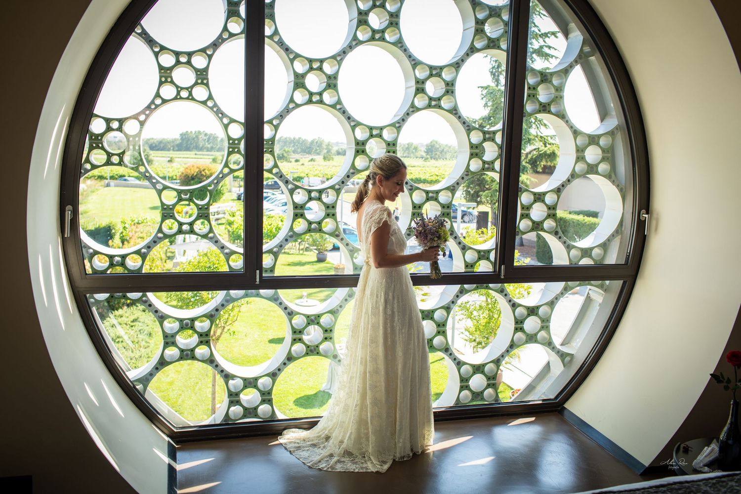 Bride in white gown stands before large circular window with geometric pattern, holding bouquet.