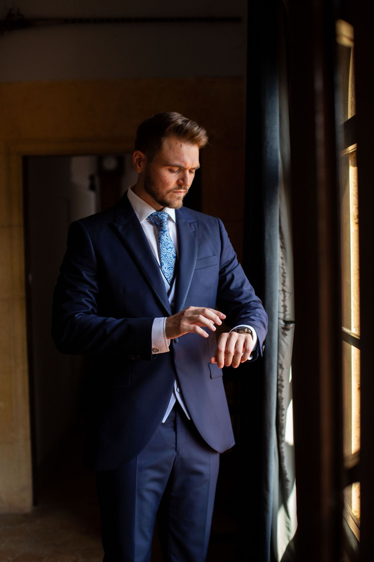 Elegant groom in navy blue suit checking his watch by a window before the wedding ceremony.