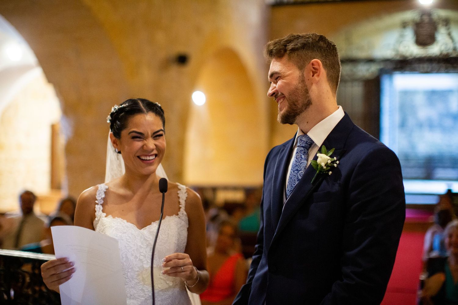 Bride and groom share a joyful laugh during wedding vows at a beautiful stone church ceremony.