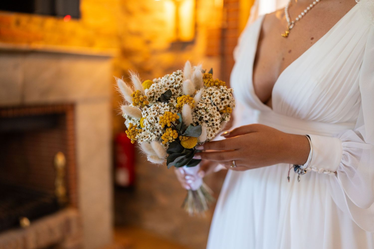 Bride in white gown holding yellow and white wildflower bouquet near fireplace.