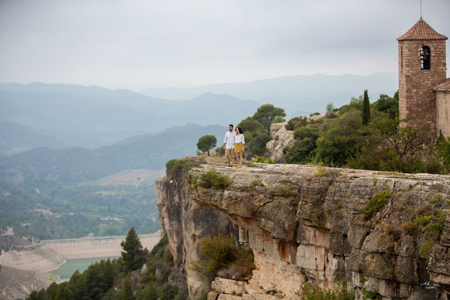 Couple standing on a dramatic cliff edge beside a medieval stone tower with sweeping mountain valley views.