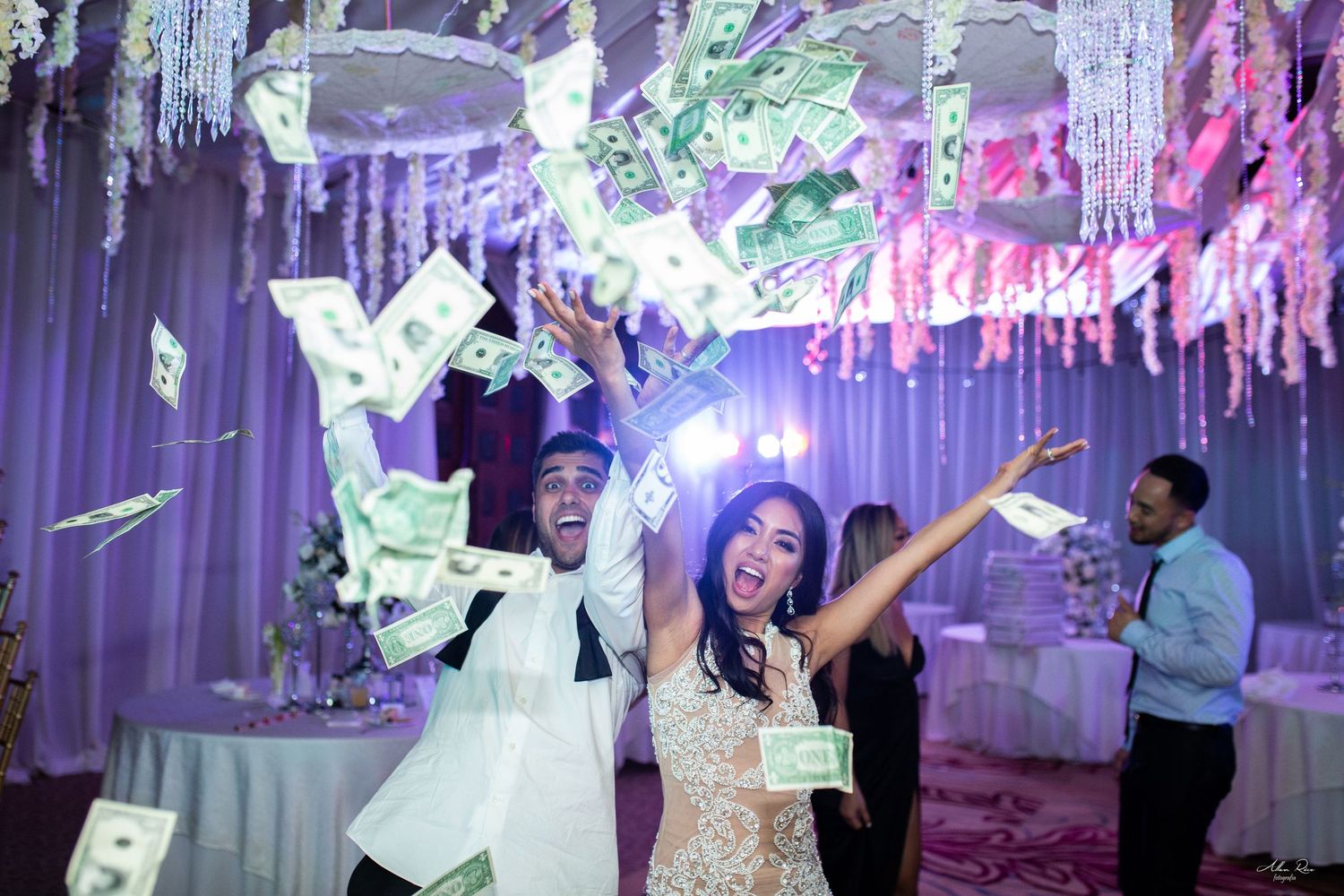Excited couple celebrating surrounded by flying dollar bills at a lavishly decorated wedding reception venue.