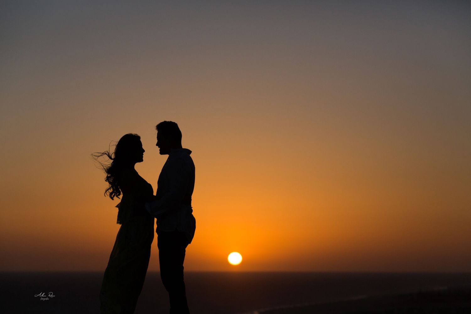 Couple silhouetted against a vibrant orange sunset sky in a romantic embrace.