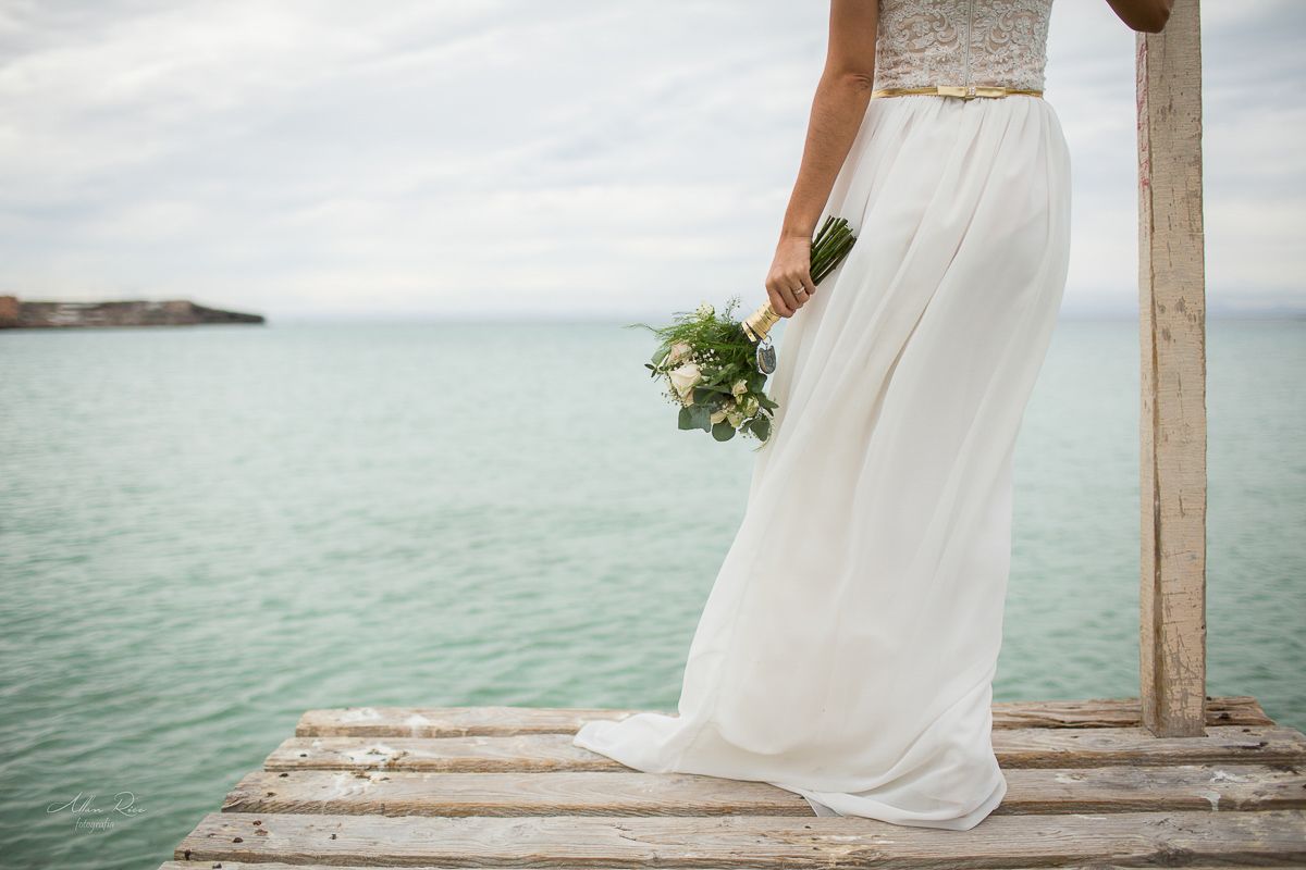 Elegant bride in white gown holding green bouquet standing on rustic pier over turquoise sea.