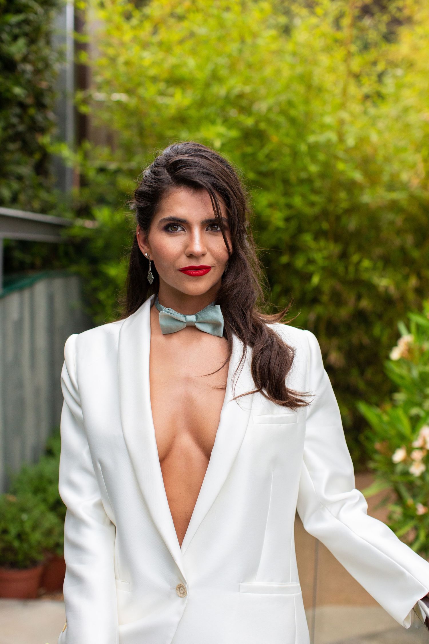 Confident bride in a deep-V white blazer suit with a sage green bow tie poses in a green outdoor garden setting.