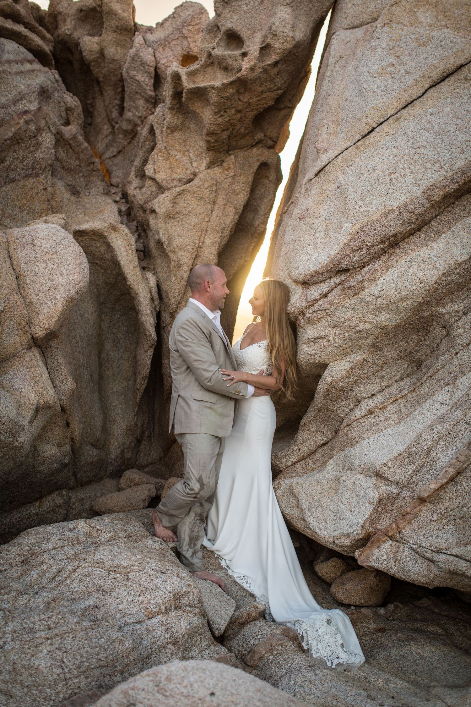 Bride and groom stand intimately between dramatic granite rock formations with sunlight glowing behind.