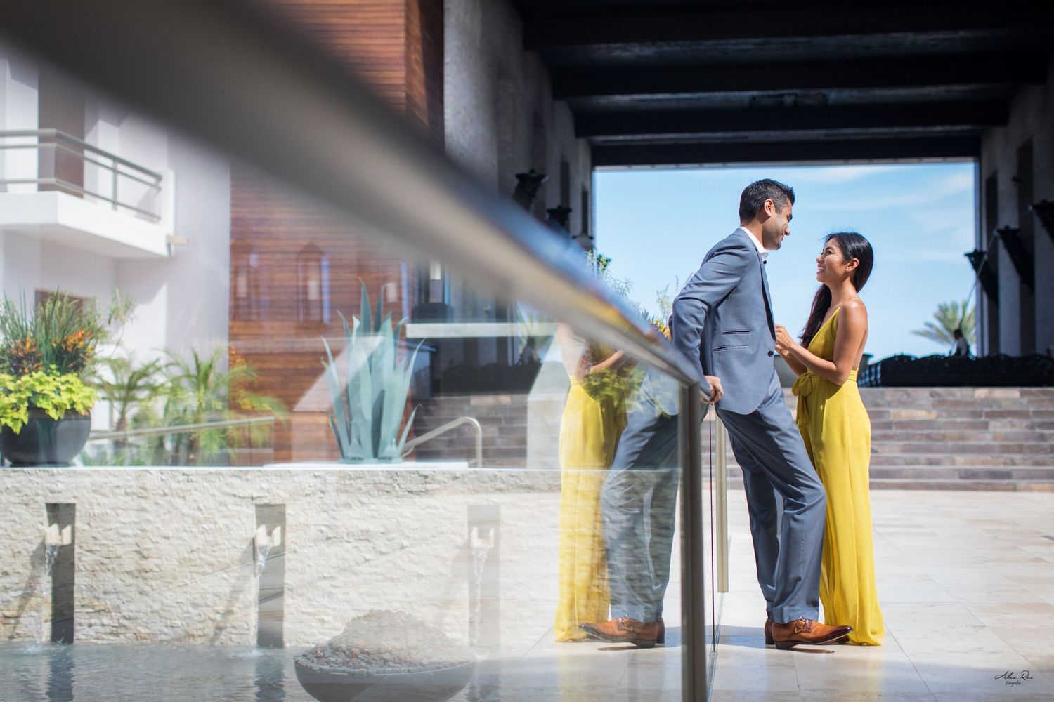 Couple in formal attire sharing a romantic moment on a modern glass balcony overlooking a resort.