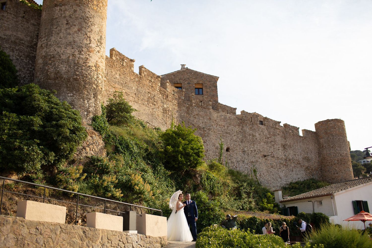 Newlyweds share a tender moment in front of a grand medieval stone castle surrounded by lush greenery.