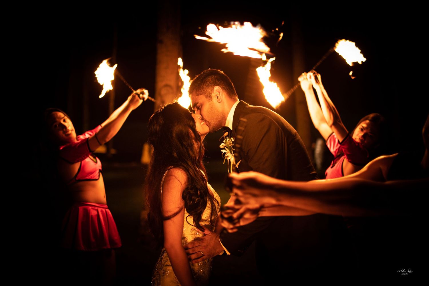 Couple shares a romantic kiss encircled by fire performers holding blazing torches at a night wedding.