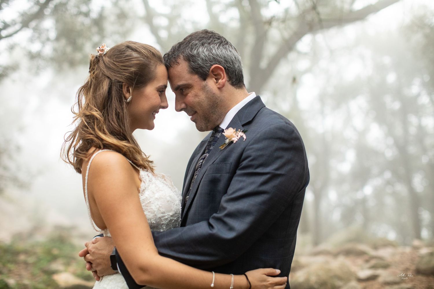 Happy bride and groom touch foreheads and smile in a misty forest wedding portrait.