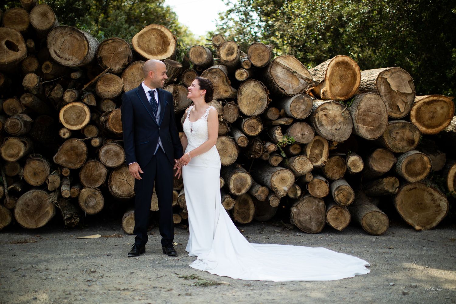 Bride and groom laughing together in front of a large stack of cut logs in natural setting.