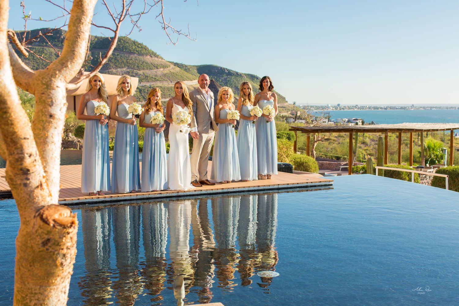 Bridesmaids in light blue gowns holding bouquets stand by a reflective infinity pool with ocean views.