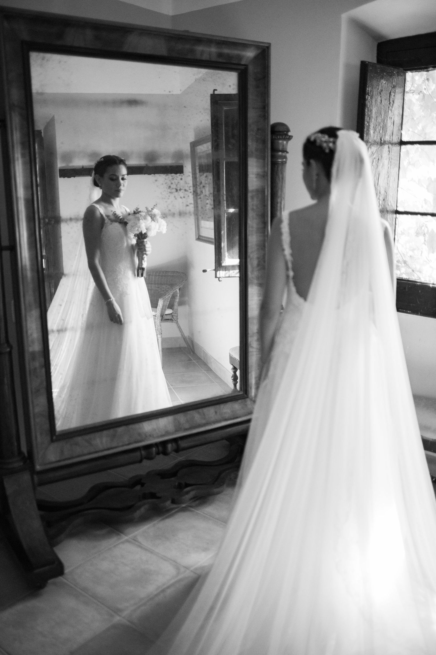 Bride in white gown and veil admires her reflection in a large ornate mirror in black and white.