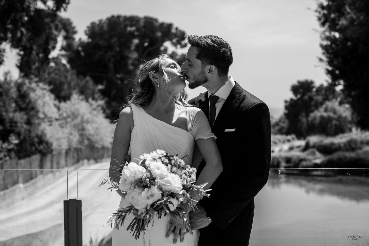 Black and white photo of a bride and groom sharing a romantic moment outdoors near a pond, bride holding a bouquet.