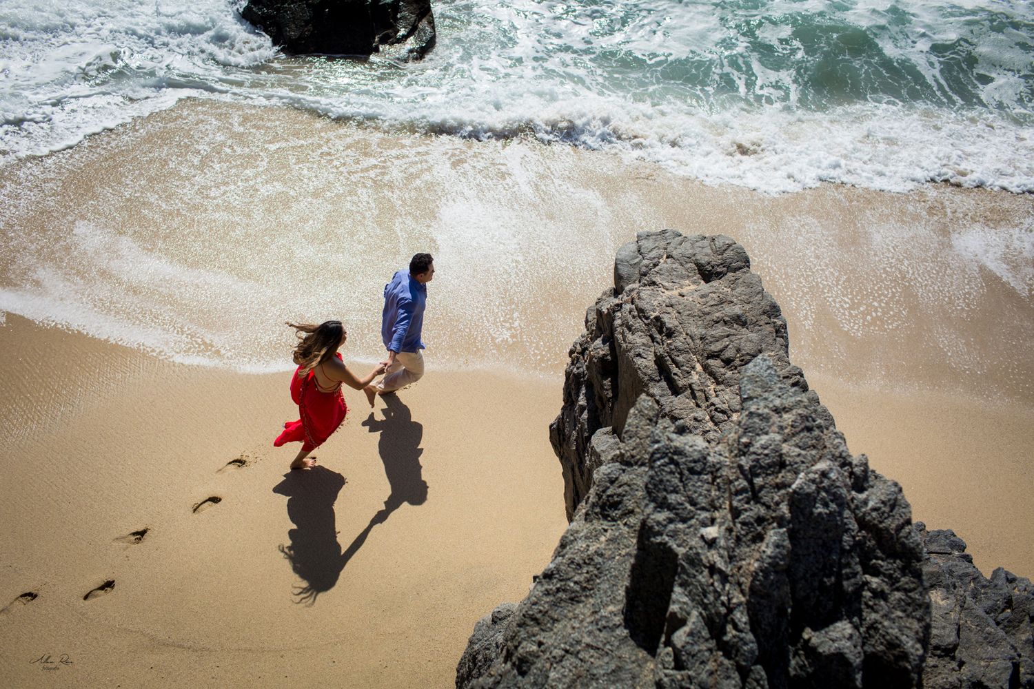 Couple holding hands walking on a sandy beach near rocky cliffs with ocean waves.