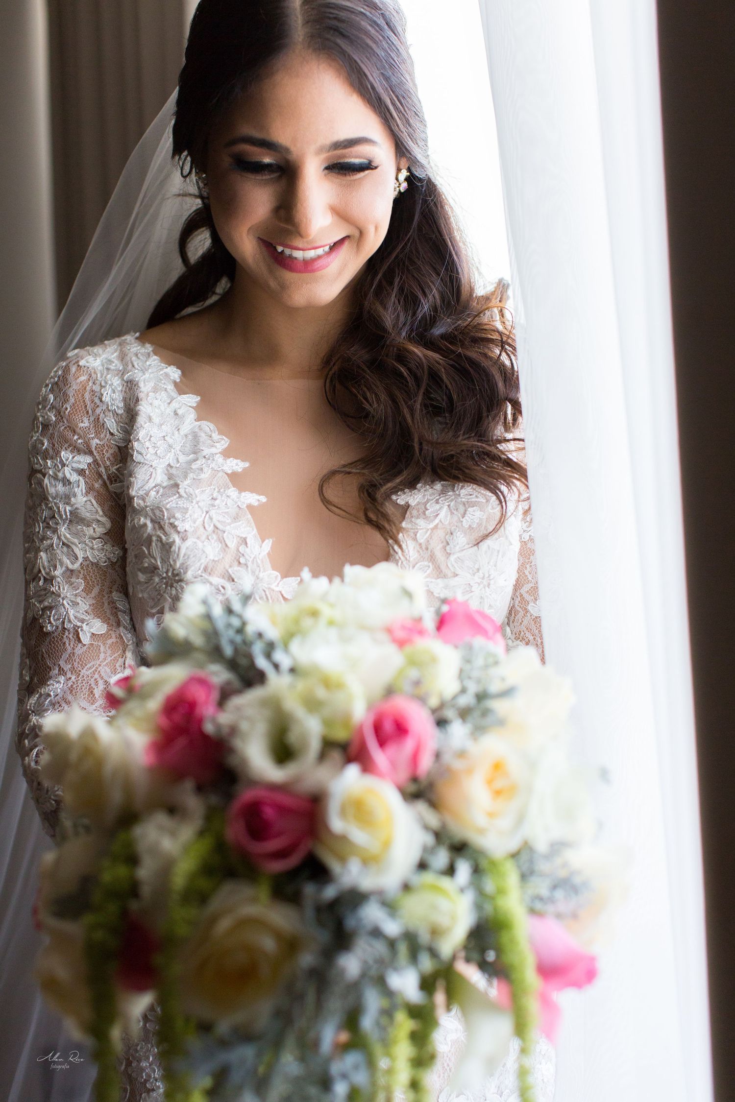Smiling bride in lace long-sleeve wedding gown holding a colorful bouquet of pink and white roses near a window.